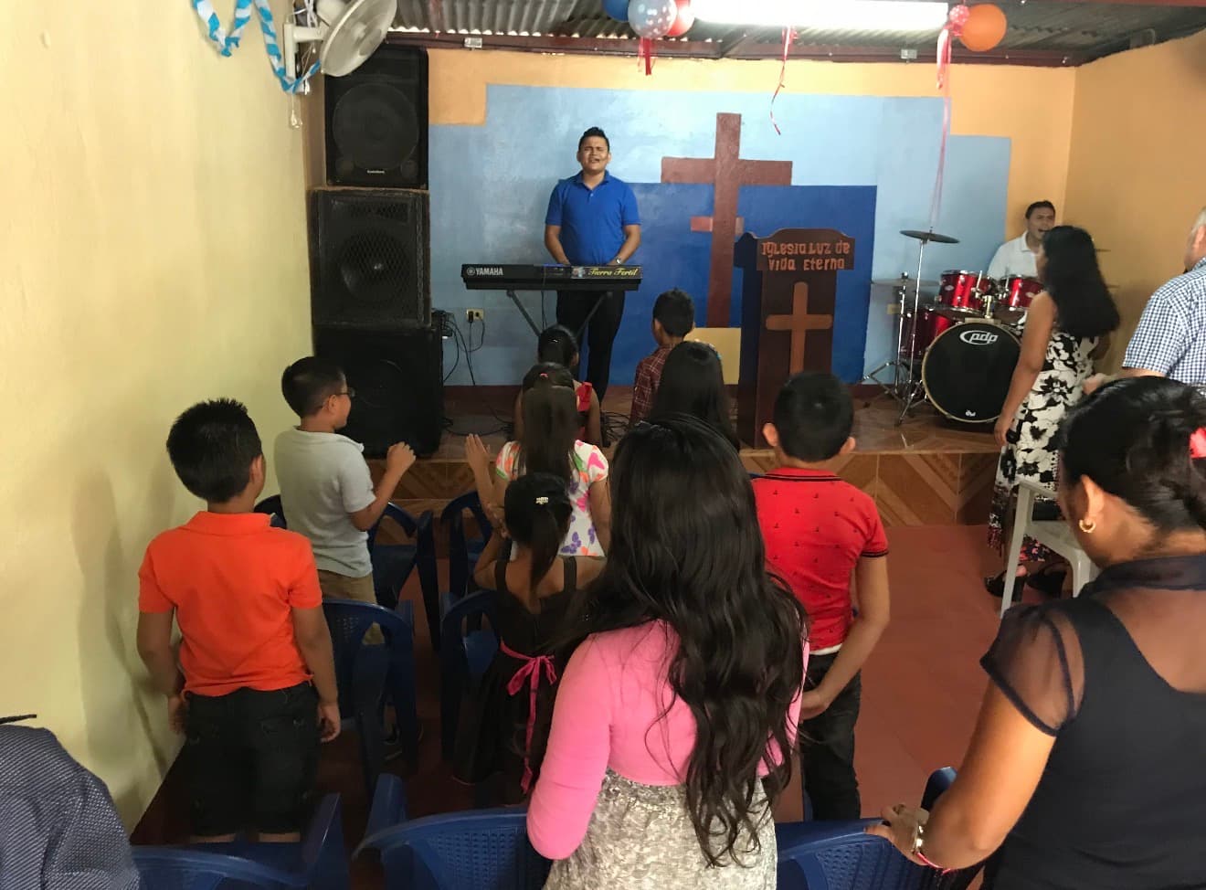 NiƱos y adultos en una iglesia pequeƱa frente a un hombre tocando el teclado.