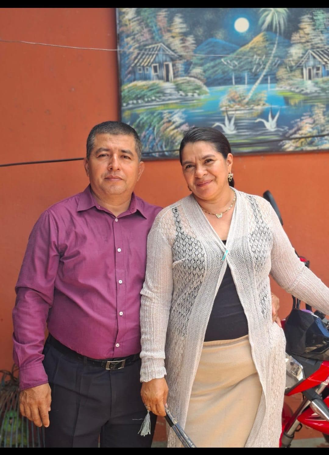 Hombre con camisa morada y mujer con cárdigan blanco posan ante una pared naranja.